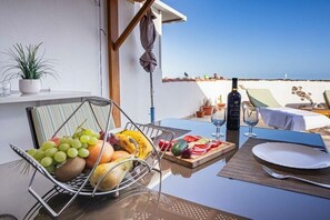 Una taula de menjador amb una cistella de fruita, una ampolla de vi i un vas de vi a un balcó amb vista al mar.