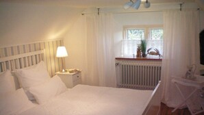 Quarto com cama, mesa de cabeceira, luminária, janela com cortinas e radiador.