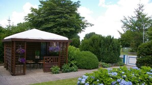 Um gazebo de madeira com telhado branco, rodeado por um jardim com arbustos de hortênsia.