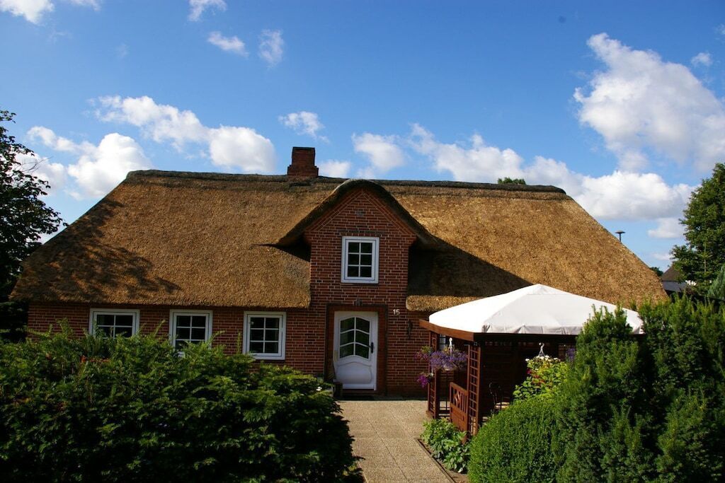 Une maison traditionnelle en briques avec un toit de chaume, une porte d’entrée et un petit porche agrémenté d’un parasol blanc.