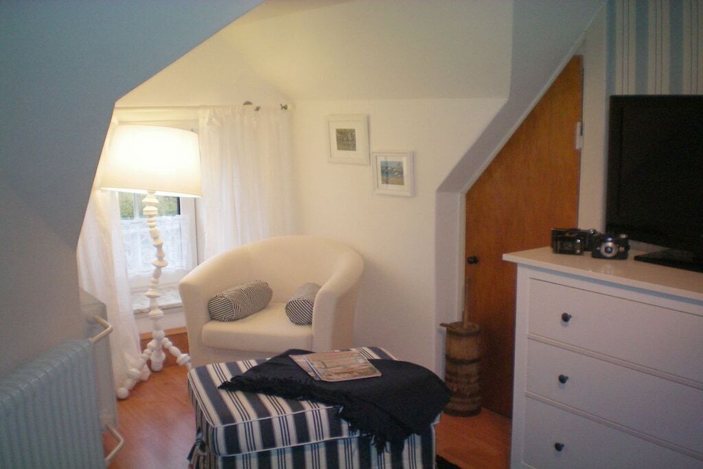 Ein gemütliches Zimmer mit einem weißen Sessel, einem gestreiften Hocker, einer Holzkommode und einem Fernseher.
