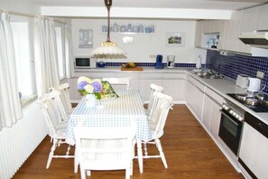 Une cuisine avec une table à manger dressée pour quatre personnes, des chaises blanches, une nappe à carreaux bleus et blancs, et un luminaire suspendu.