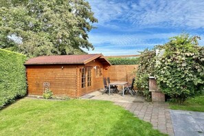 Een houten chalet met een terras van baksteen, omgeven door een verzorgd gazon en een haag.
