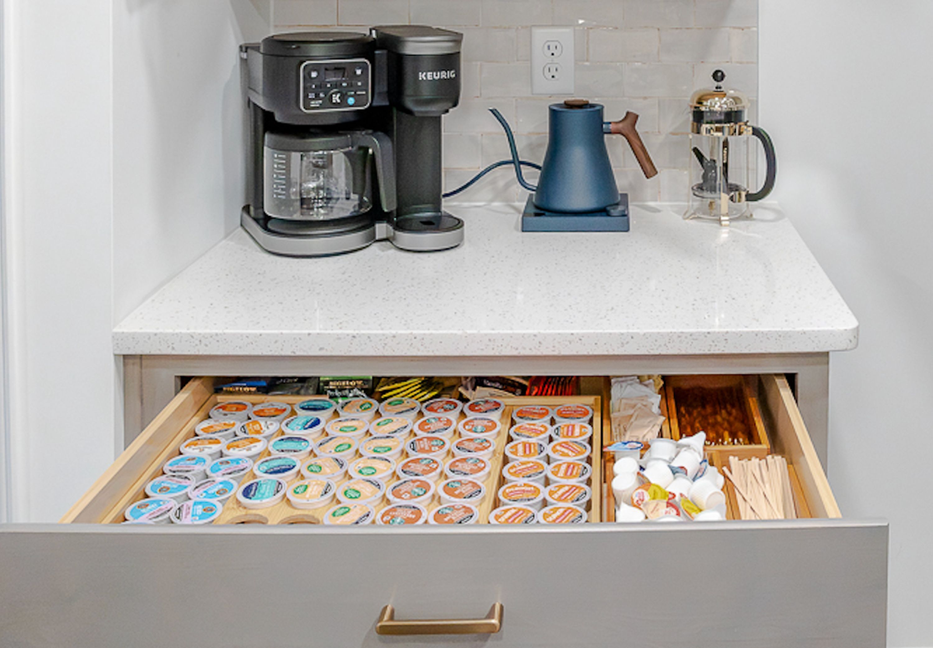A kitchen counter with a coffee maker, a kettle, and a coffee pot. Below, a drawer contains various food items.