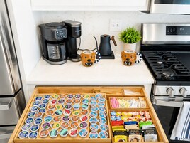 Una cocina con cafetera, hervidor y estufa. Abajo, un cajón contiene diversos alimentos y recipientes.