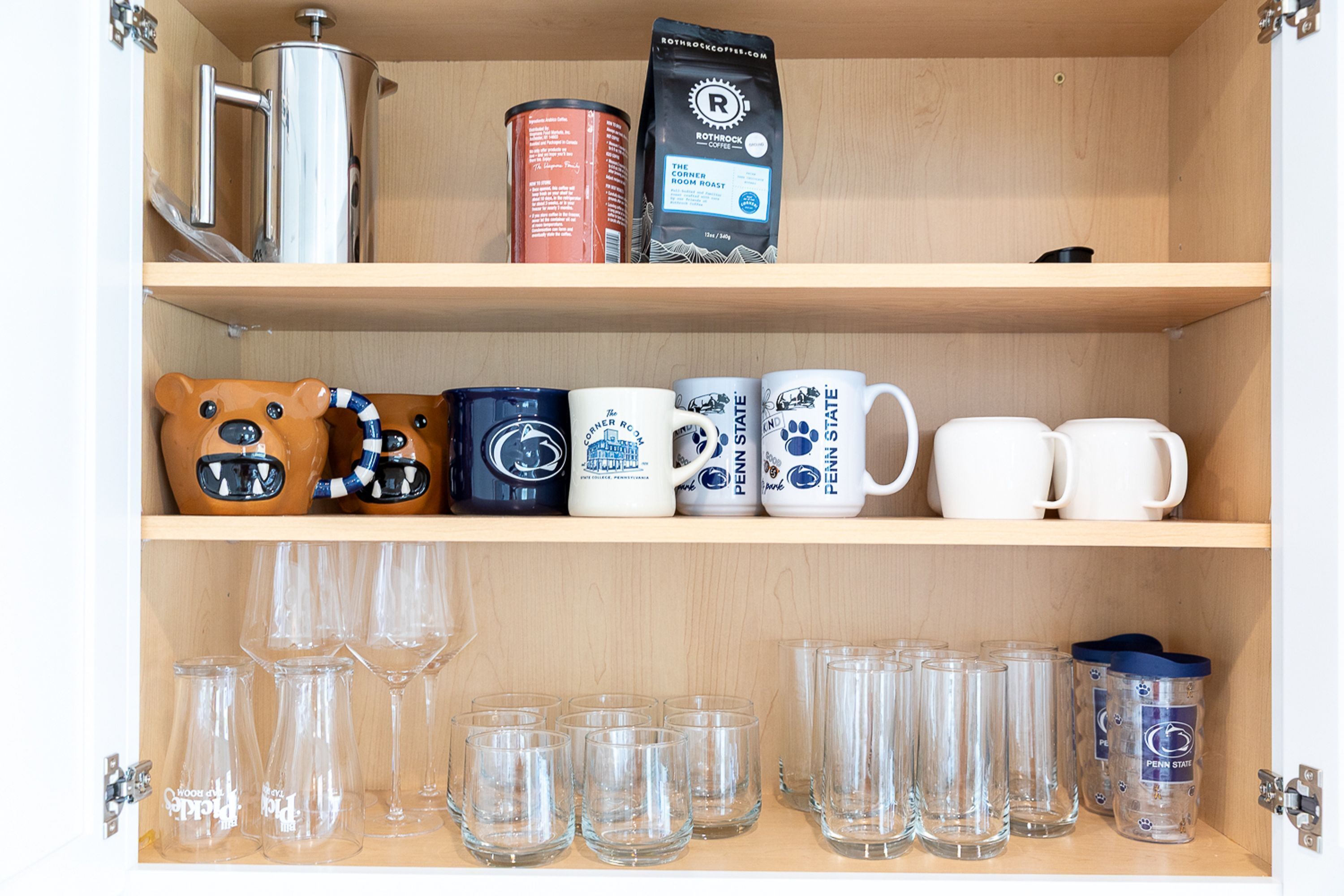 A wooden cabinet with shelves containing mugs, glasses, a coffee maker, and a canister.