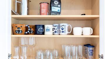A wooden cabinet with shelves containing mugs, glasses, a coffee maker, and a canister.