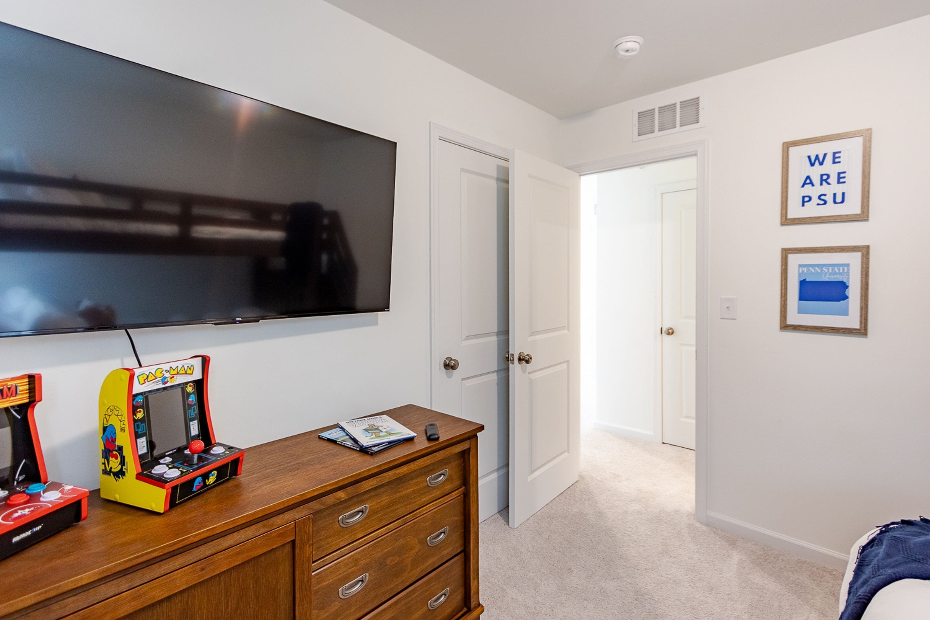 A bedroom with a TV mounted on the wall, a wooden dresser with drawers, a Pac-Man arcade game, and framed pictures on the wall.