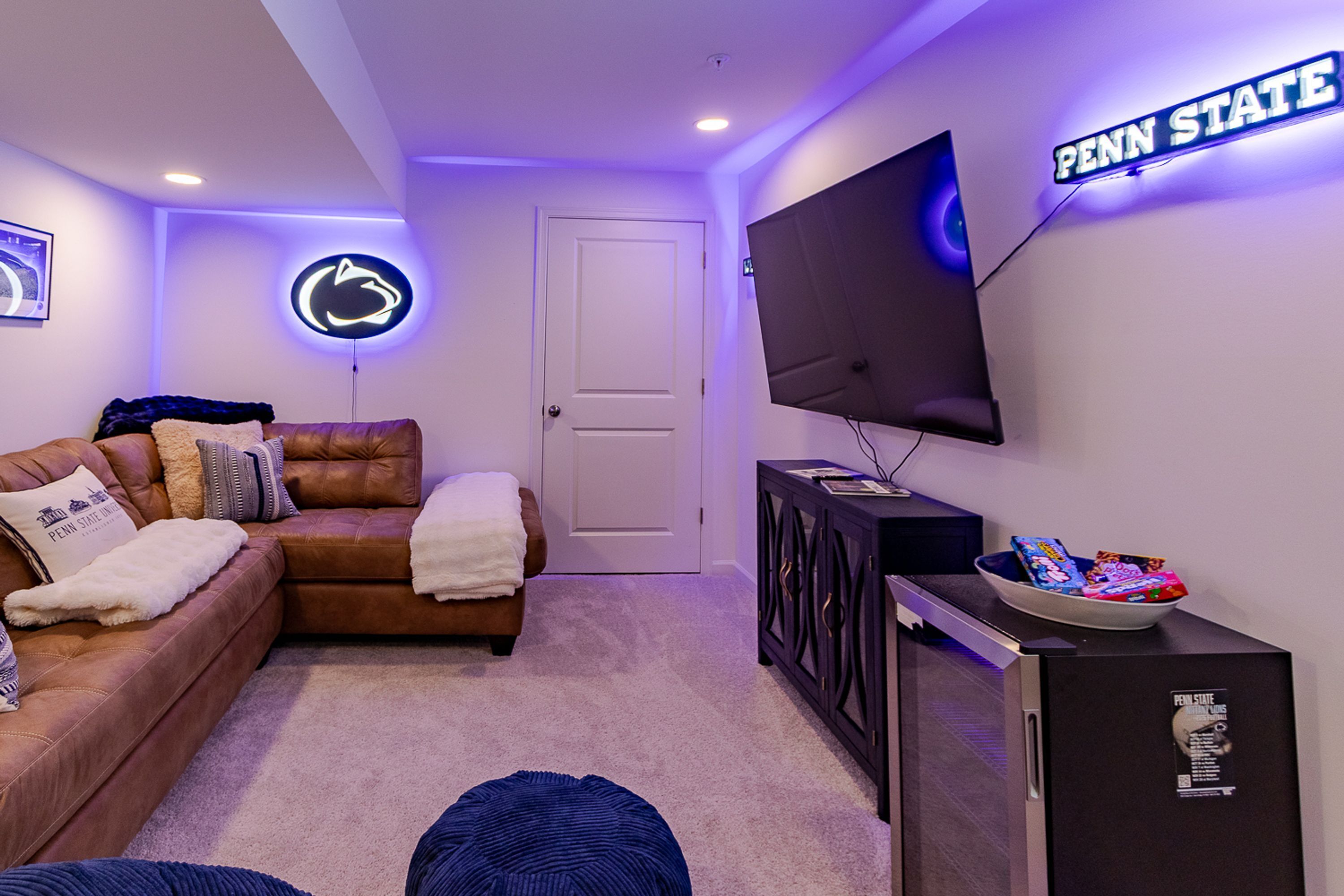 A living room with a large flat-screen TV mounted on the wall, a leather sofa, a small cabinet, and a neon sign.