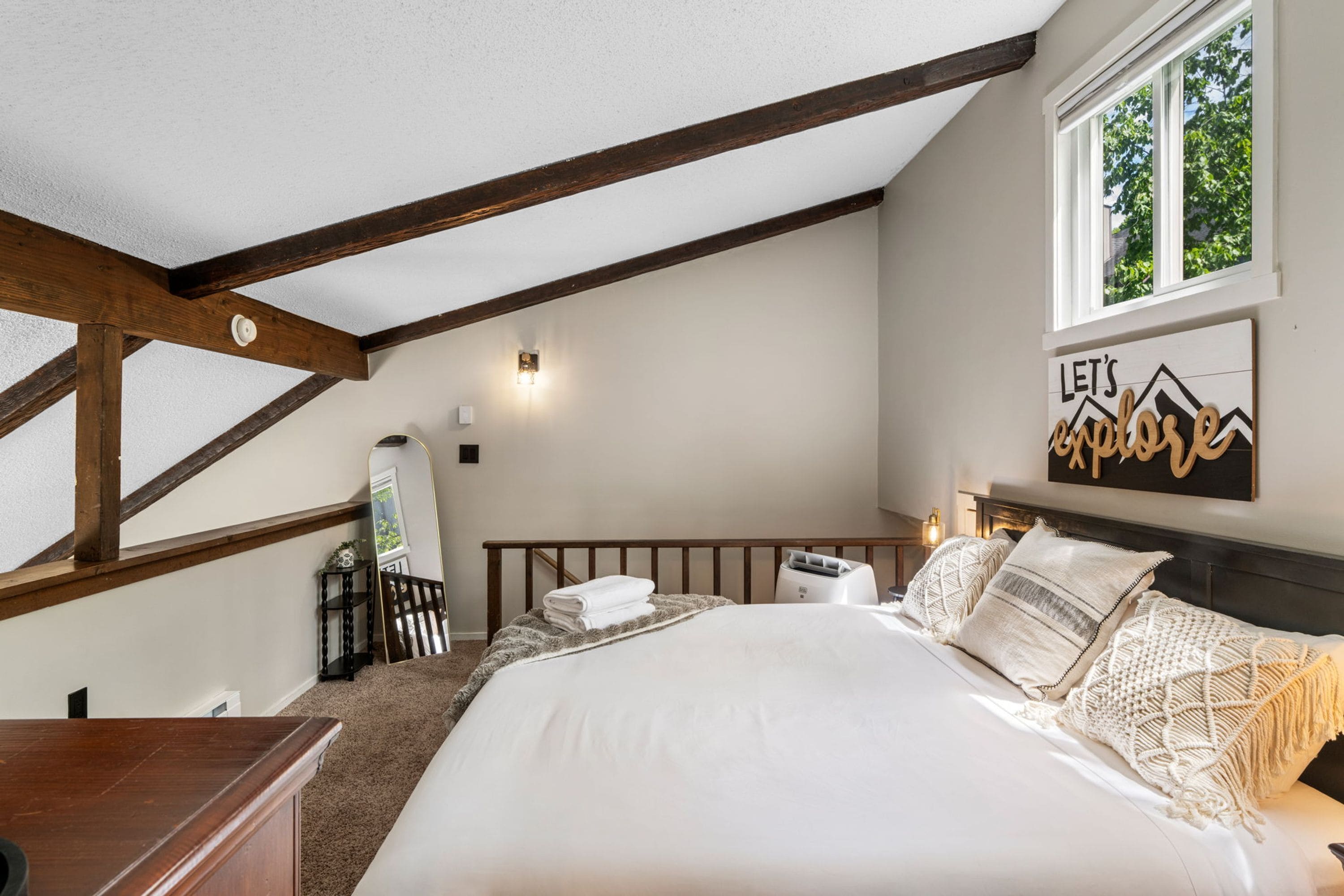 A bedroom with a large bed, a window, a wall-mounted artwork, and a wooden ceiling beam structure.