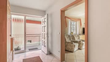 Un couloir mène à un espace de vie comprenant un canapé et des fauteuils, une porte-fenêtre coulissante et un sol carrelé.