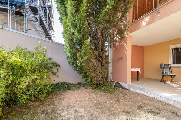 A residential building with a balcony, a small garden area with a tree, and a patio with a table and chair.