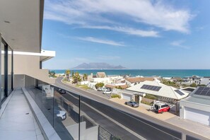 A balcony with a view of a coastal street, a mountain in the distance, and a clear sky. - The Blouberg Lookout (Cape Town)