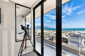 A balcony with a telescope, a clear view of the ocean and a distant mountain, and a residential area in the background. - The Blouberg Lookout (Cape Town)