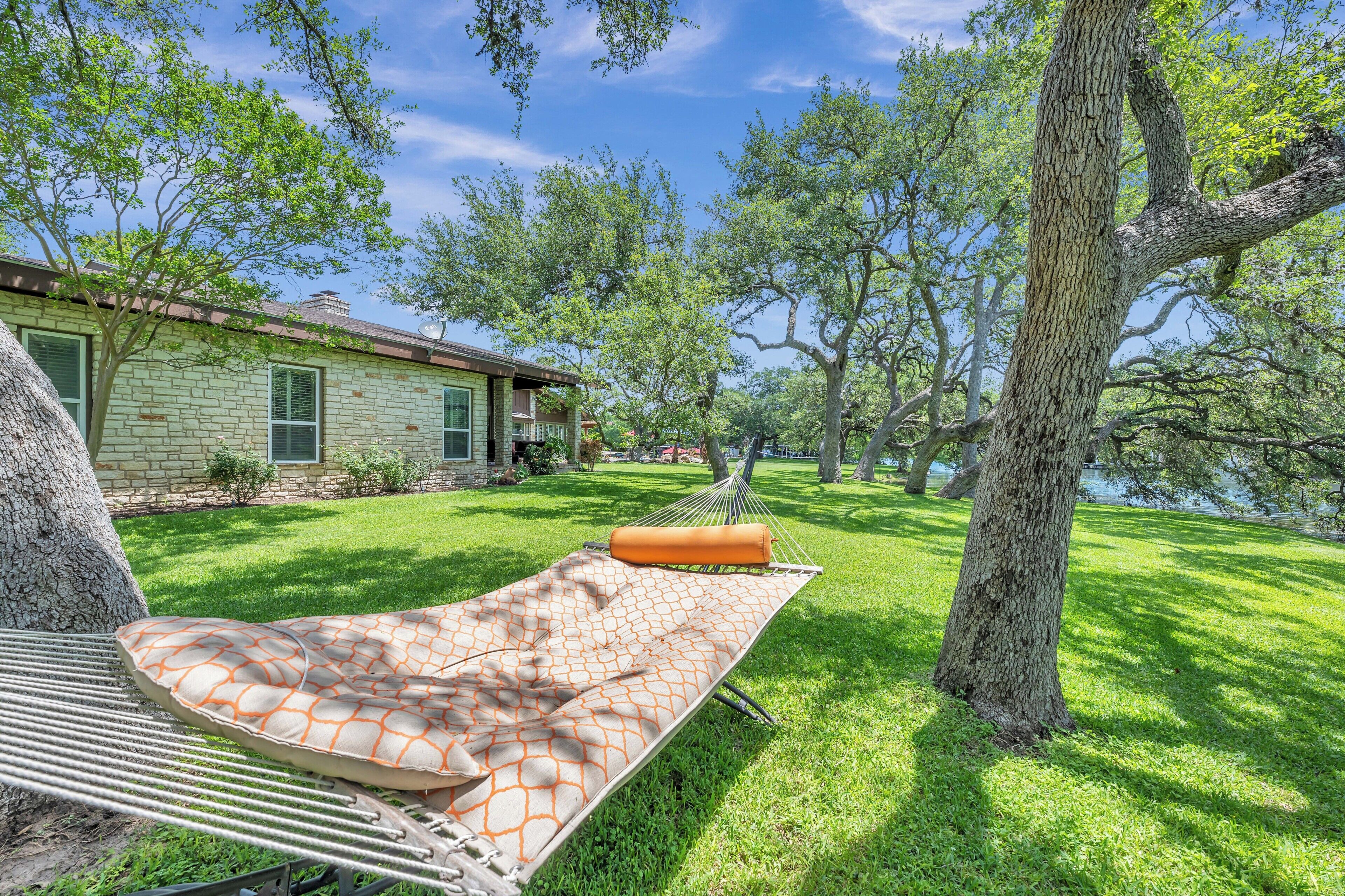 A hammock with a patterned cushion in a grassy yard with a stone building and trees.