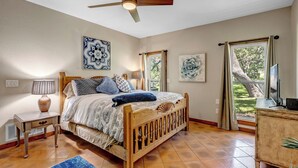 Ein Schlafzimmer mit einem hölzernen Bett, einem Deckenventilator, Kunstwerken an den Wänden und einem Fenster mit Blick auf Grünflächen.