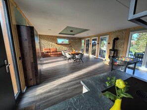 A modern living room with a stone wall, wooden flooring, a sofa, a dining table with chairs, and a foosball table. - An oasis of peace, where charm and elegance meet nature—a chalet in Western Lusatia (Haselbachtal)