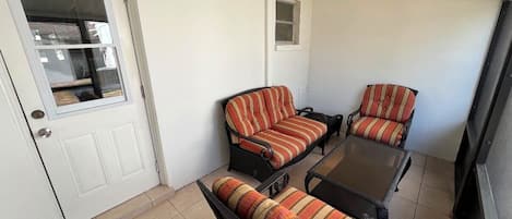 Una habitación con una puerta blanca, un ventanal pequeño y dos sofás de mimbre negro con cojines a rayas.