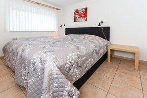 Een slaapkamer met een bed, een nachtkastje, een bankje, een radiator, een raam met gordijnen en een aan de muur hangend kunstwerk.