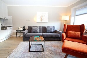Una sala de estar moderna con un sofá, una mesa de centro y una silla. Hay una zona de cocina con armarios y una encimera. Una lámpara y un cuadro están en la pared.