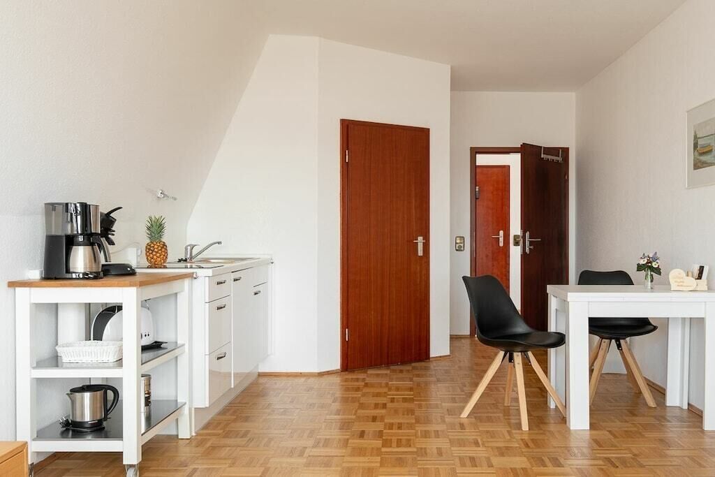 Una cocina compacta con cafetera, una piña y un lavamanos. Hay un comedor con una mesa blanca y sillas negras.