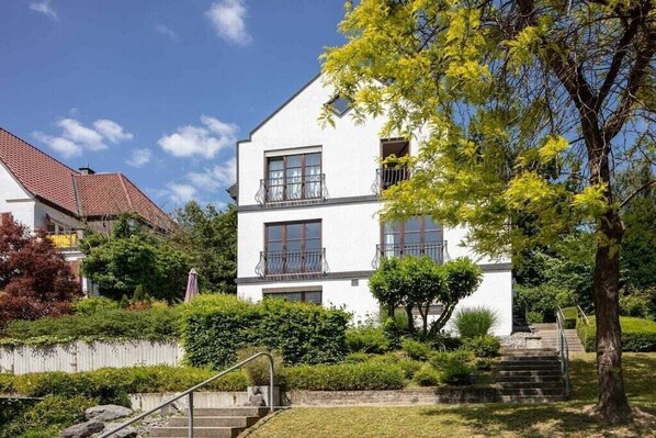 Ein zweistöckiges Gebäude mit weißer Fassade, einem Balkon im zweiten Stockwerk und einer Treppe zum Eingang.