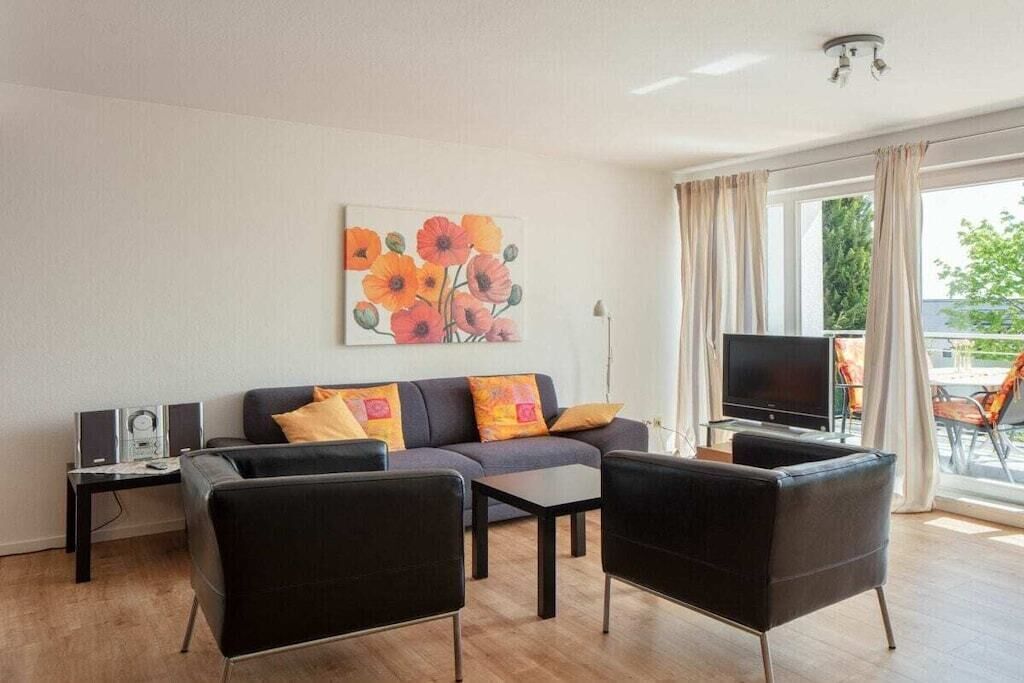 Ein modernes Wohnzimmer mit einer Couch, zwei Sesseln, einem Couchtisch, einem Flachbildfernseher und einem Blumenbild an der Wand.
