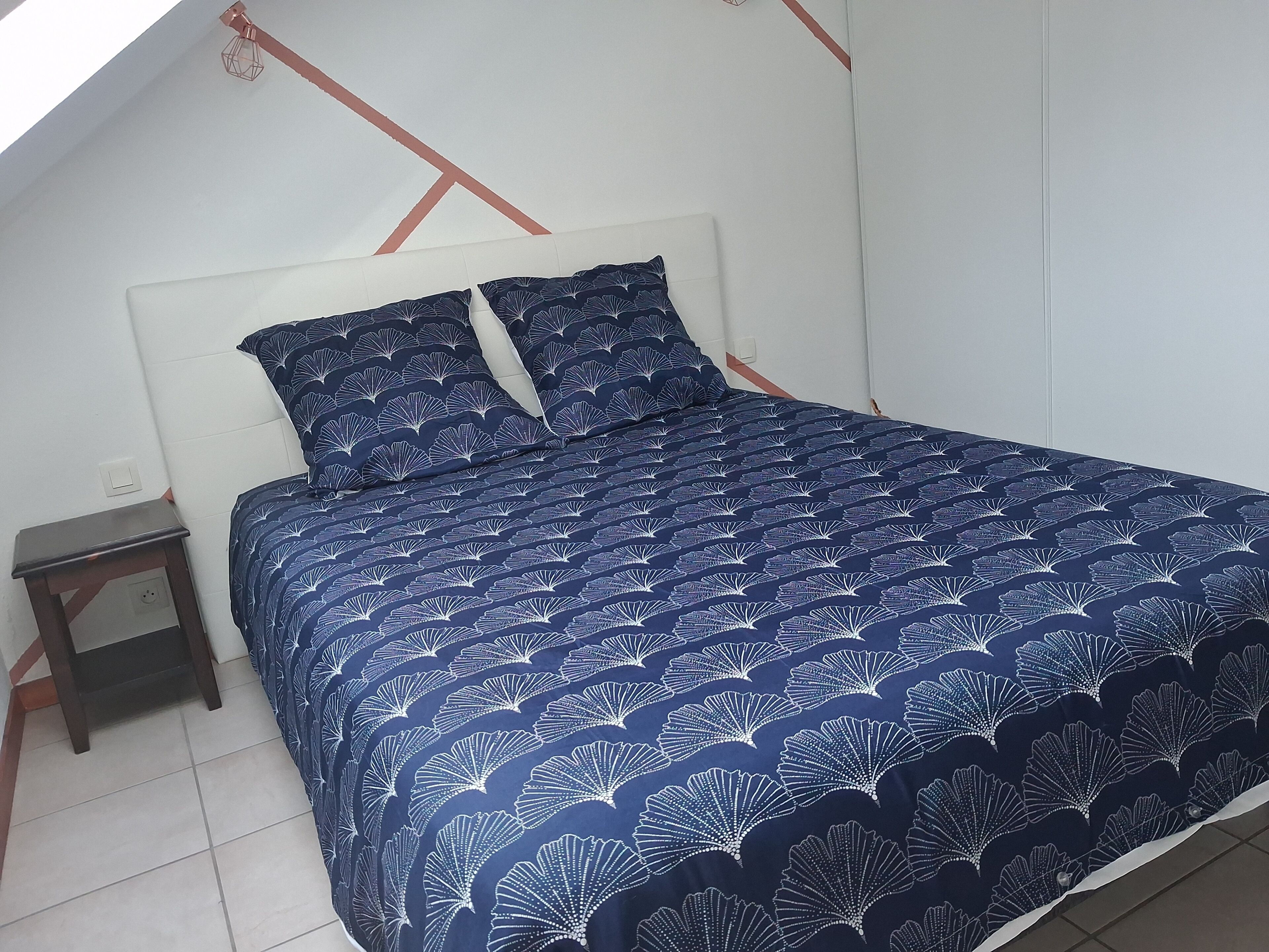 Een bed met een donkerblauwe bedspreide en passende kussens, een klein houten nachtkastje en een wit hoofdbord.