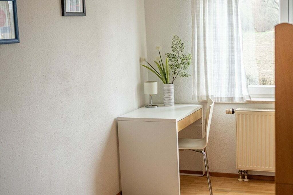 Un escritorio blanco con una silla, un jarrón con flores, una lámpara y una ventana con cortinas.