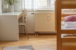 Ein kleines Zimmer mit Schreibtisch, Stuhl, Etagenbett, Heizkörper und Fenster.