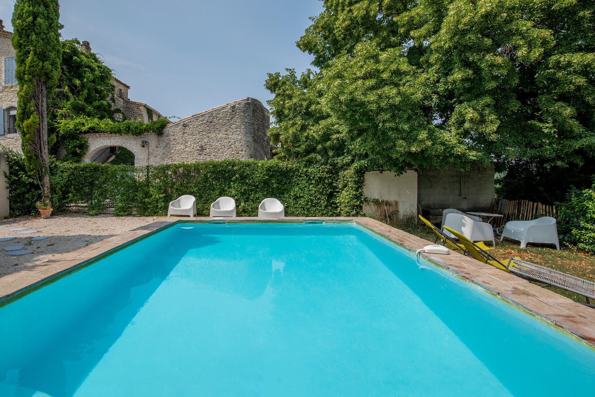 Una piscina rectangular con borde de piedra, rodeada de follaje y un edificio de piedra.