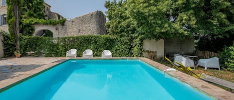 Una piscina rectangular con borde de piedra, rodeada de follaje y un edificio de piedra.