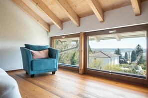 A room with a wooden ceiling, a large window with a view of a house and trees, and a blue armchair. - Holiday apartment with 1 bedroom (Überlingen)