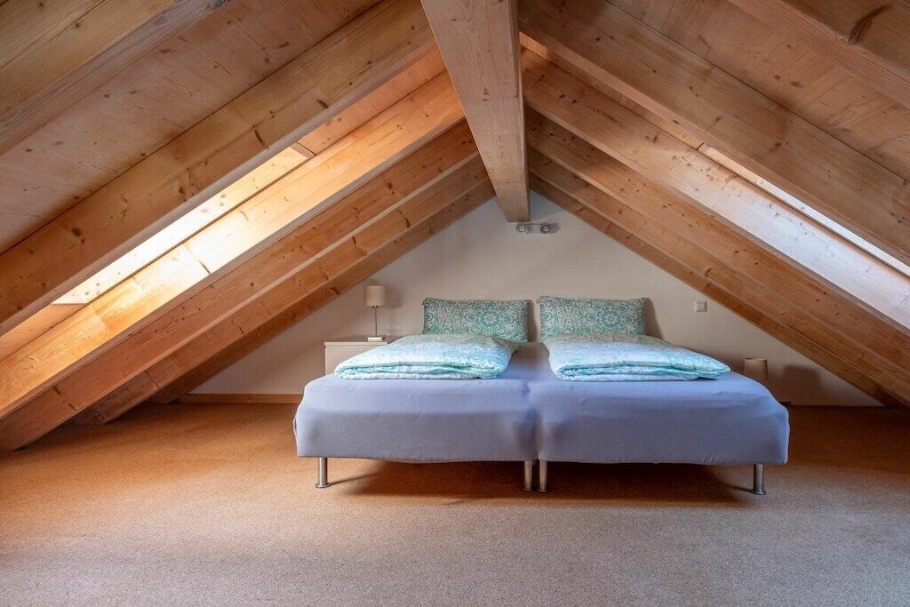 Une chambre avec un plafond en pente, un lit avec un matelas blanc et une literie bleue, et une table de chevet avec une lampe.