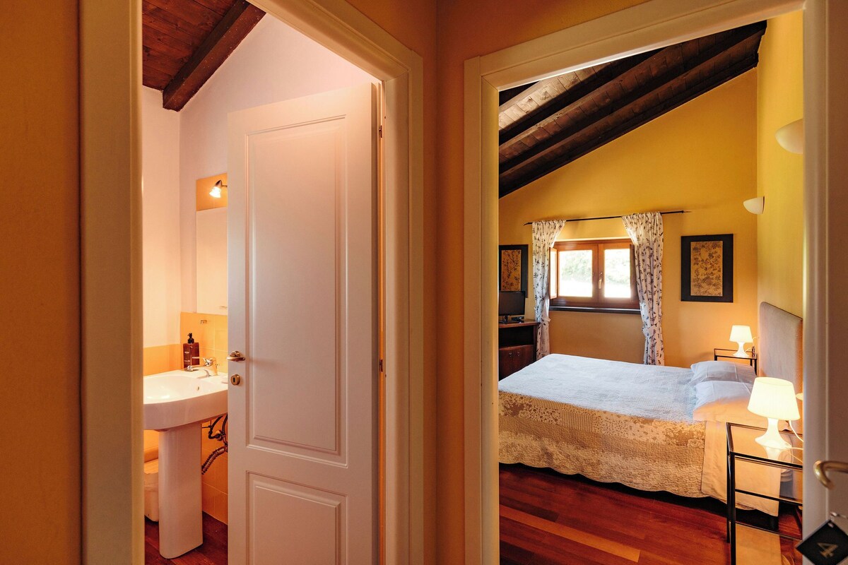 A bedroom with a bed, a nightstand, a lamp, a window with curtains, and a door leading to a bathroom.