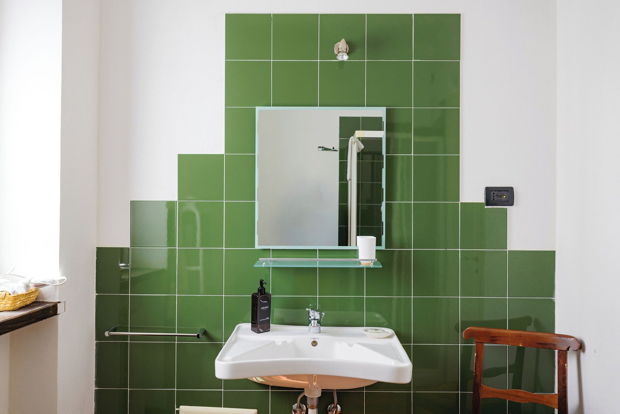 Une salle de bain avec un lavabo blanc, des carreaux verts, un miroir et une chaise en bois.