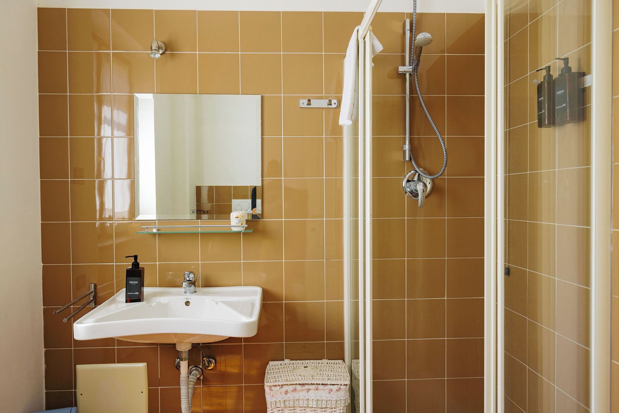 Casa de banho com uma pia branca, espelho, chuveiro com chuveiro manual e paredes revestidas a azulejos castanhos.