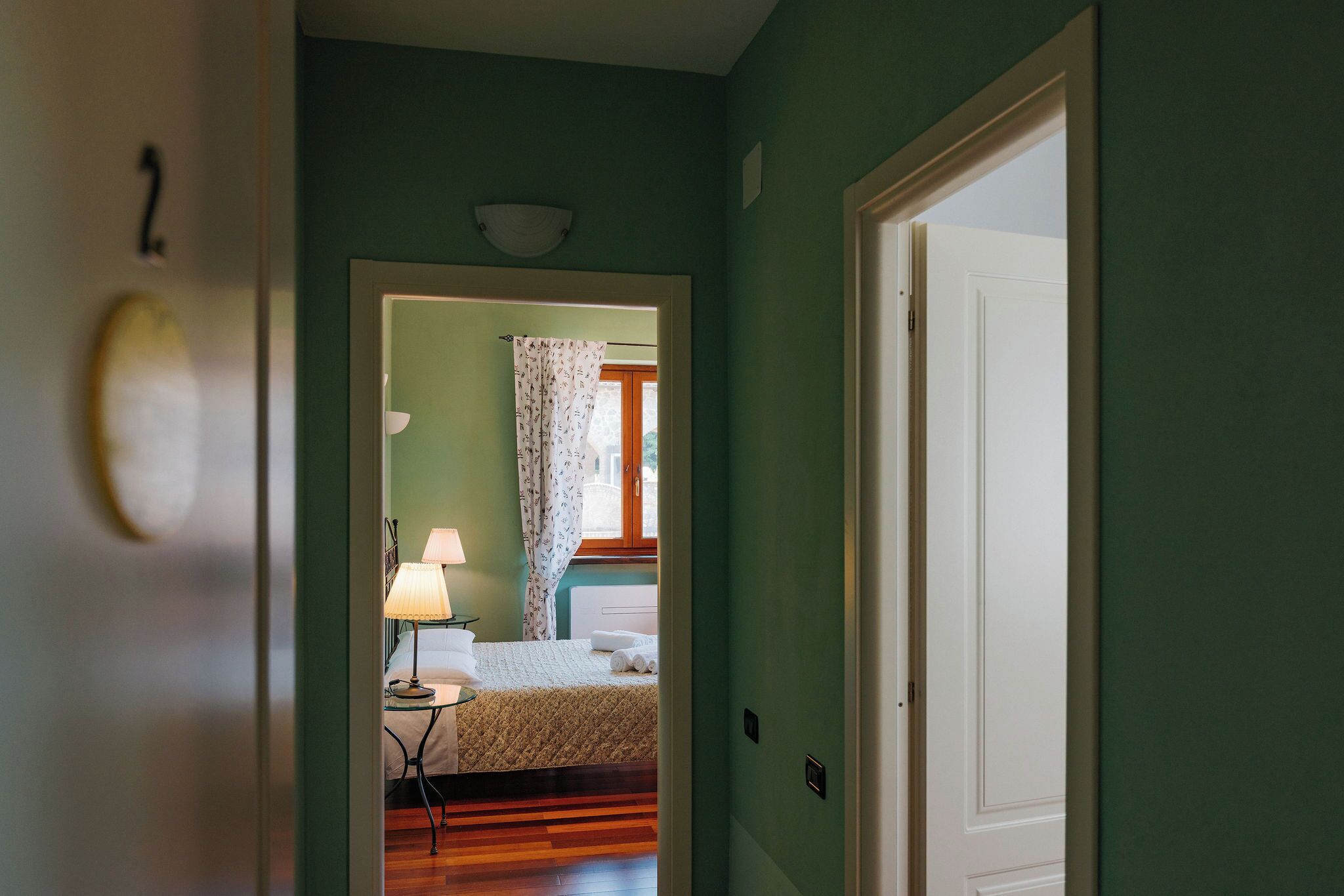 A hallway leading to a bedroom with a bed, a nightstand, a lamp, and a window with curtains.