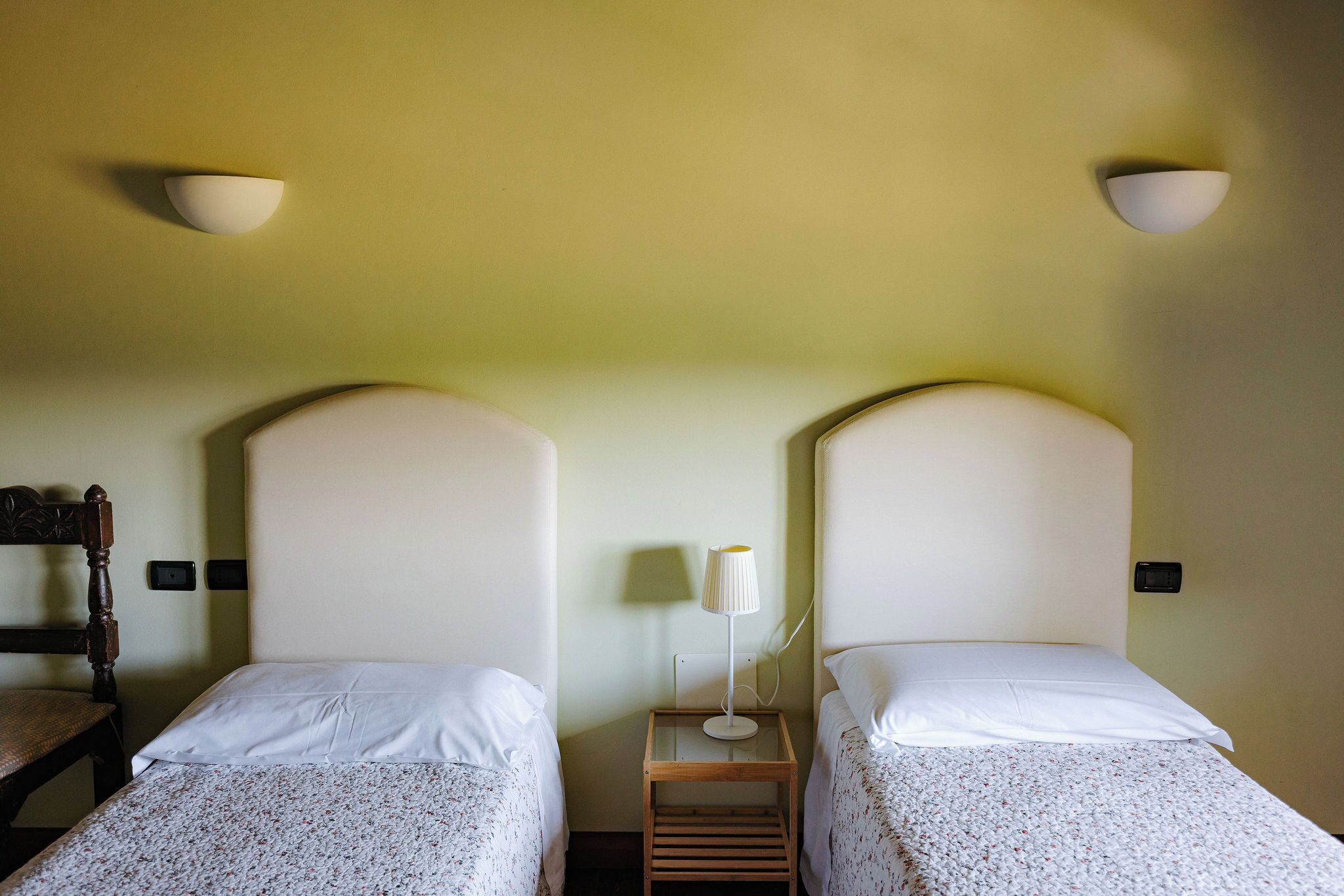 Two single beds with white linens, a wooden nightstand with a lamp, and a chair.