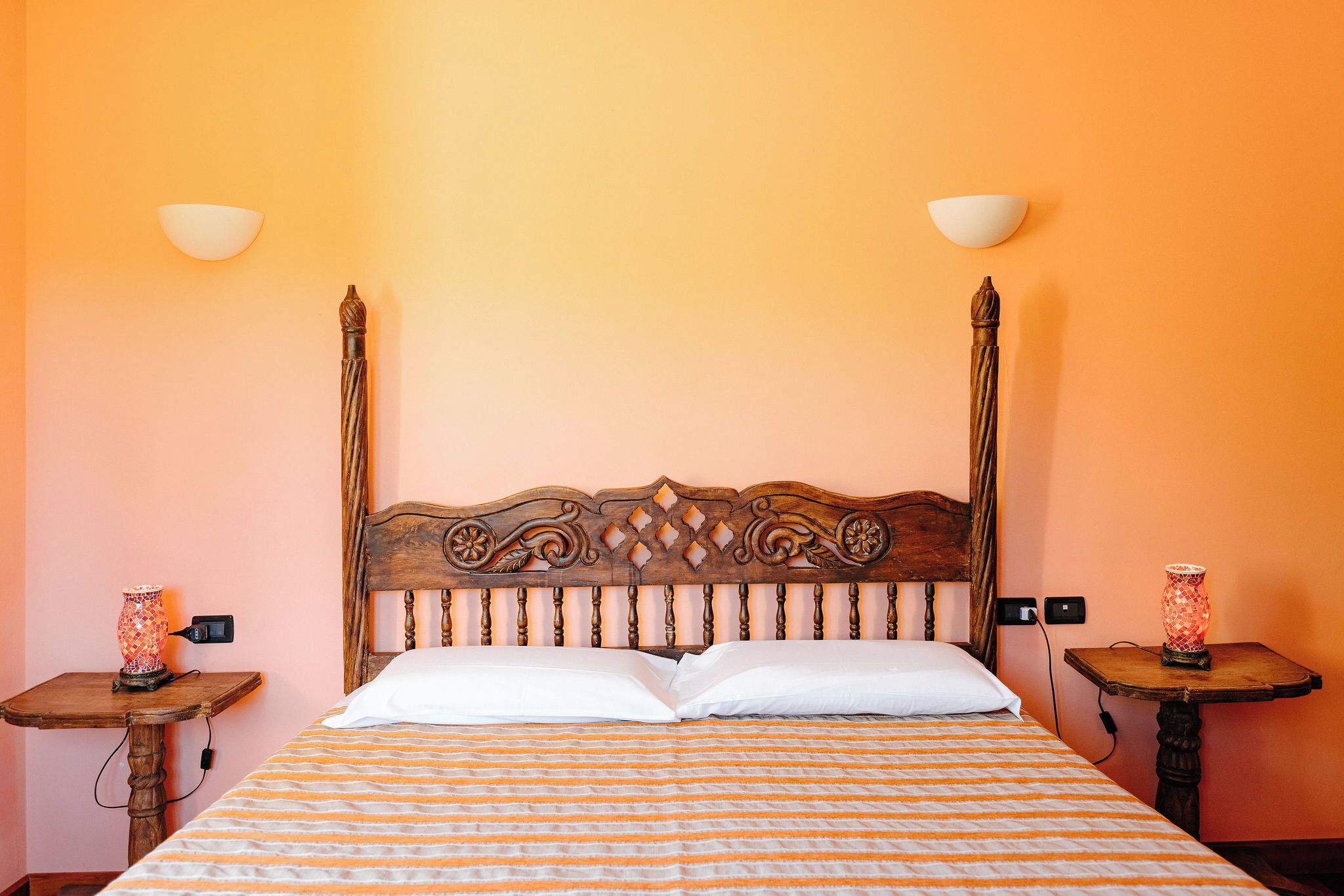 A room with a wooden headboard, two bedside tables with lamps, a bed with striped bedding, and a plain wall.
