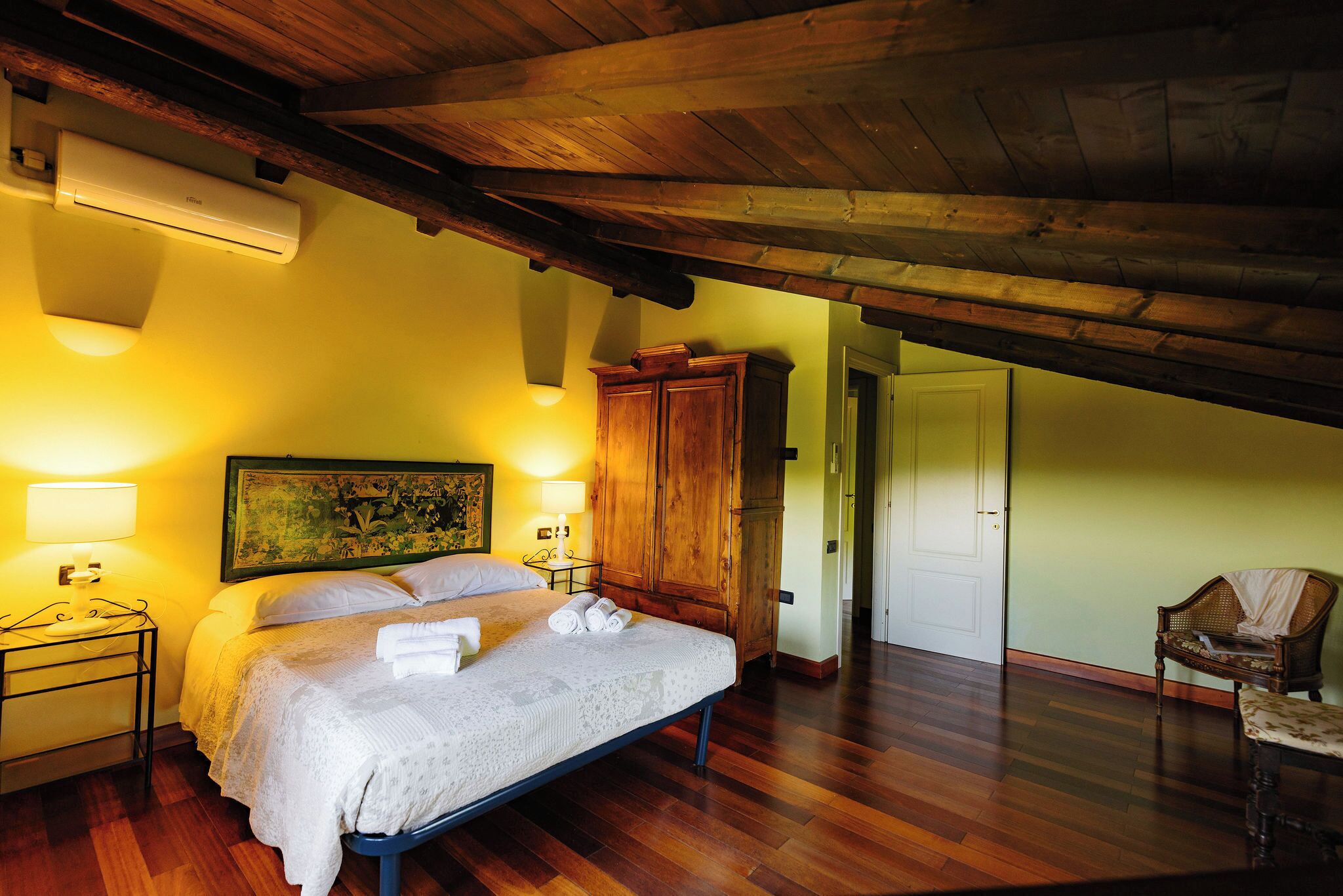 Un dormitorio con una cama, lámparas de noche, un armario de madera, una silla y un cuadro en la pared.