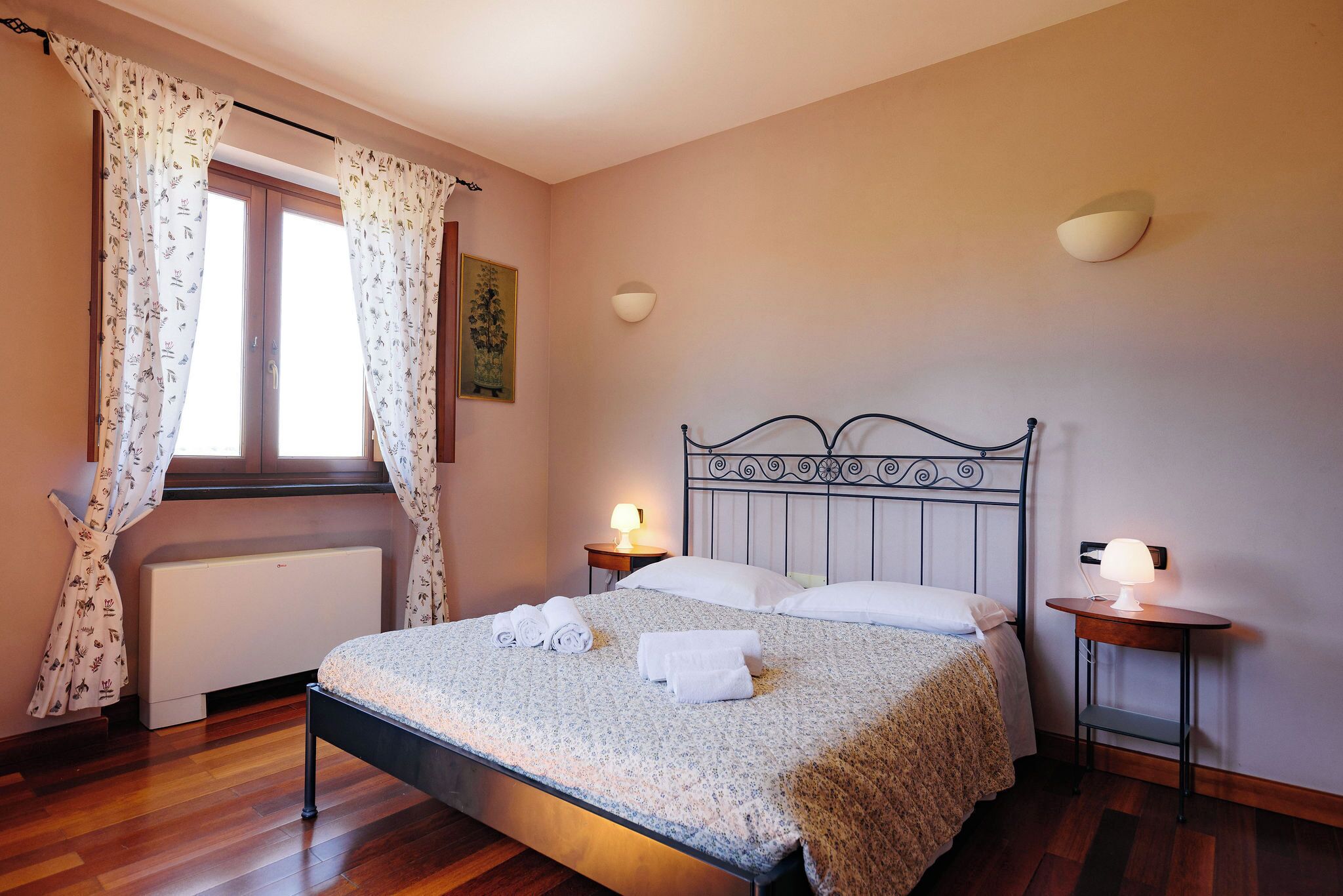 A neatly made bed with rolled towels, a window with curtains, a wall-mounted light, and a radiator.