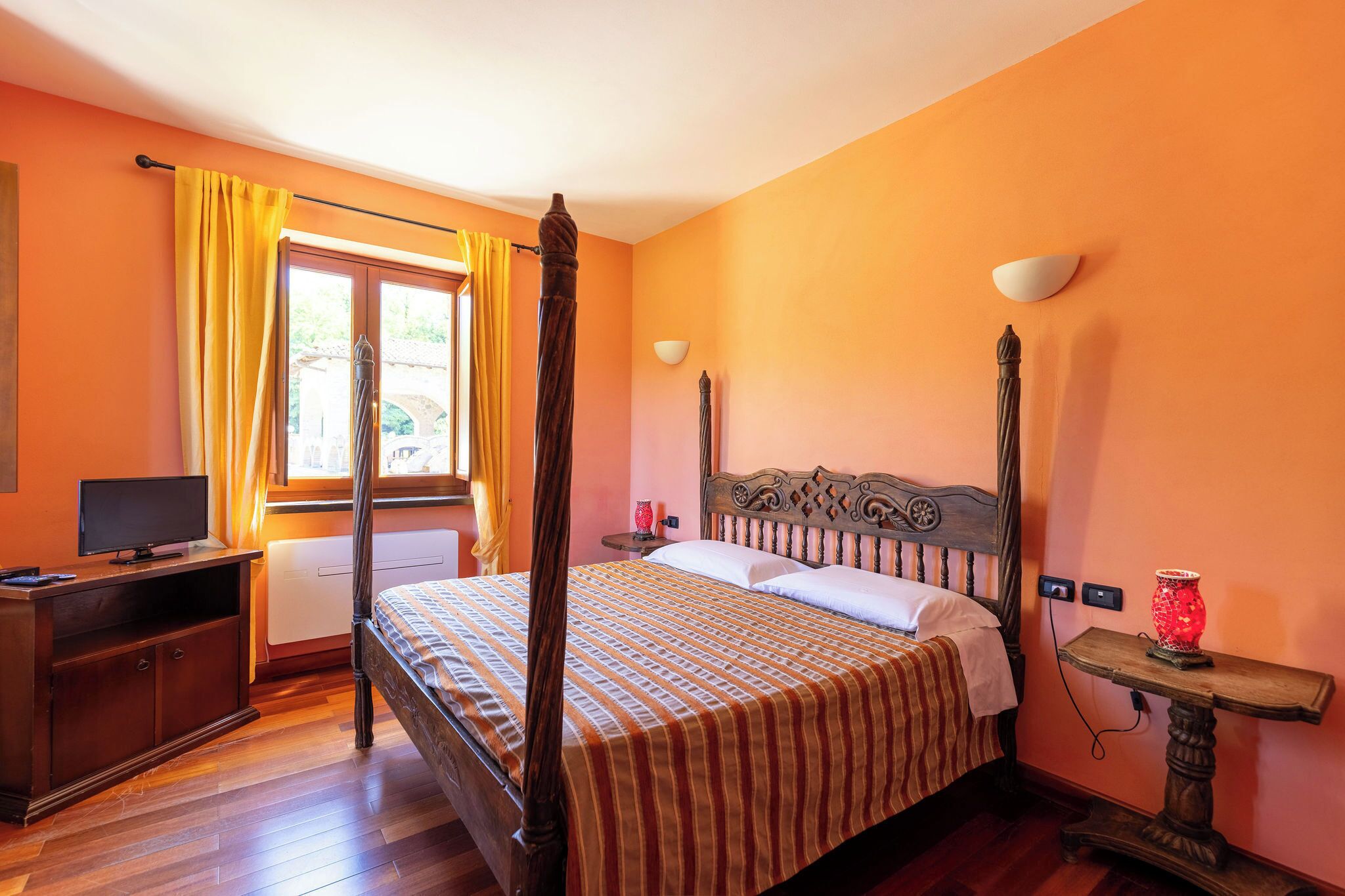 A bedroom with a four-poster bed, a wooden desk with a television, and a window with curtains.
