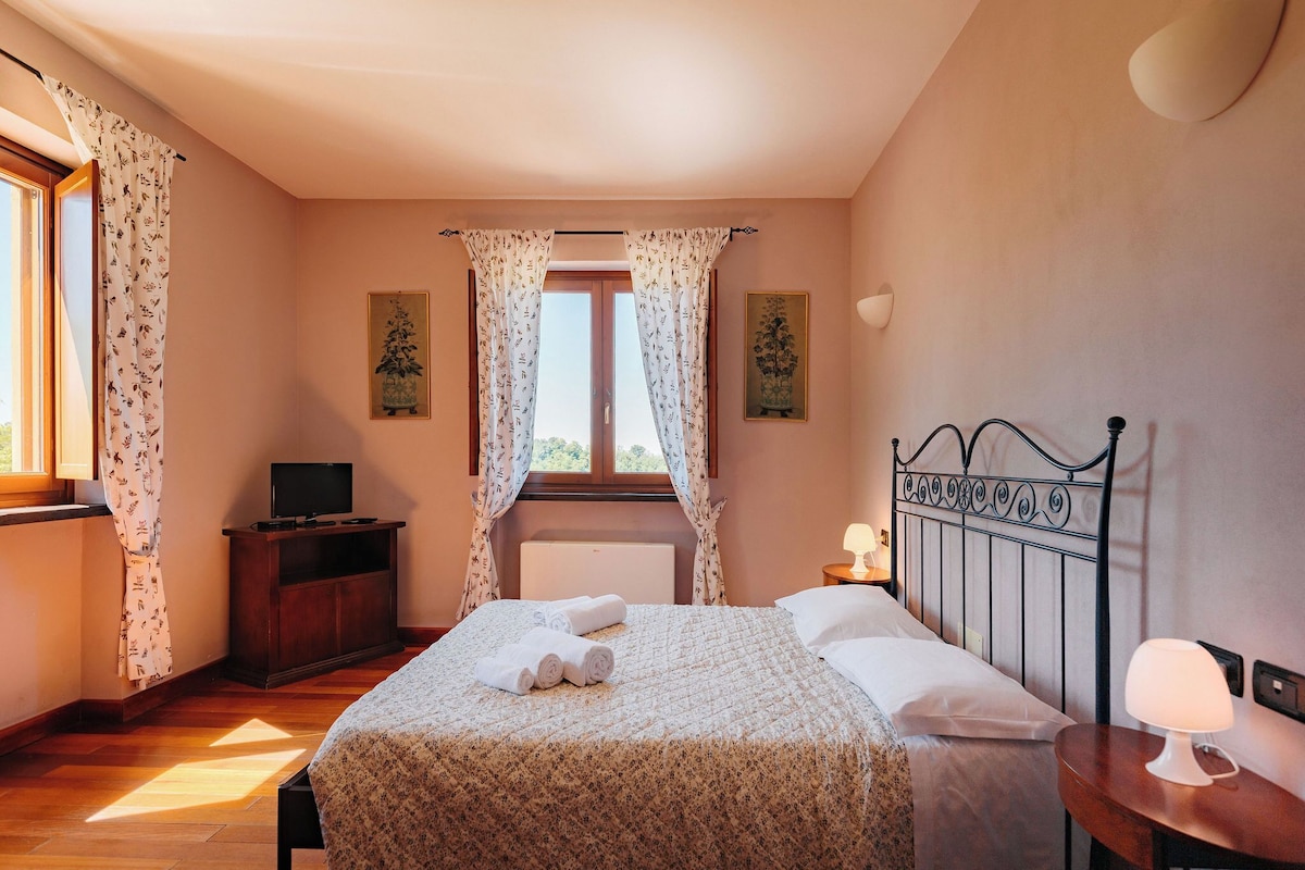 A bedroom with a bed, a bedside table with a lamp, a TV on a wooden cabinet, and a window with curtains.