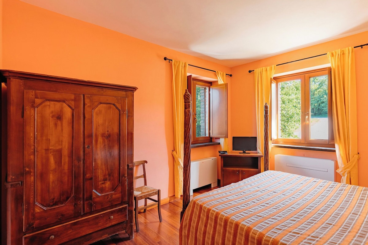 A room with a wooden wardrobe, a bed with striped bedding, a desk with a computer, and a window with curtains.
