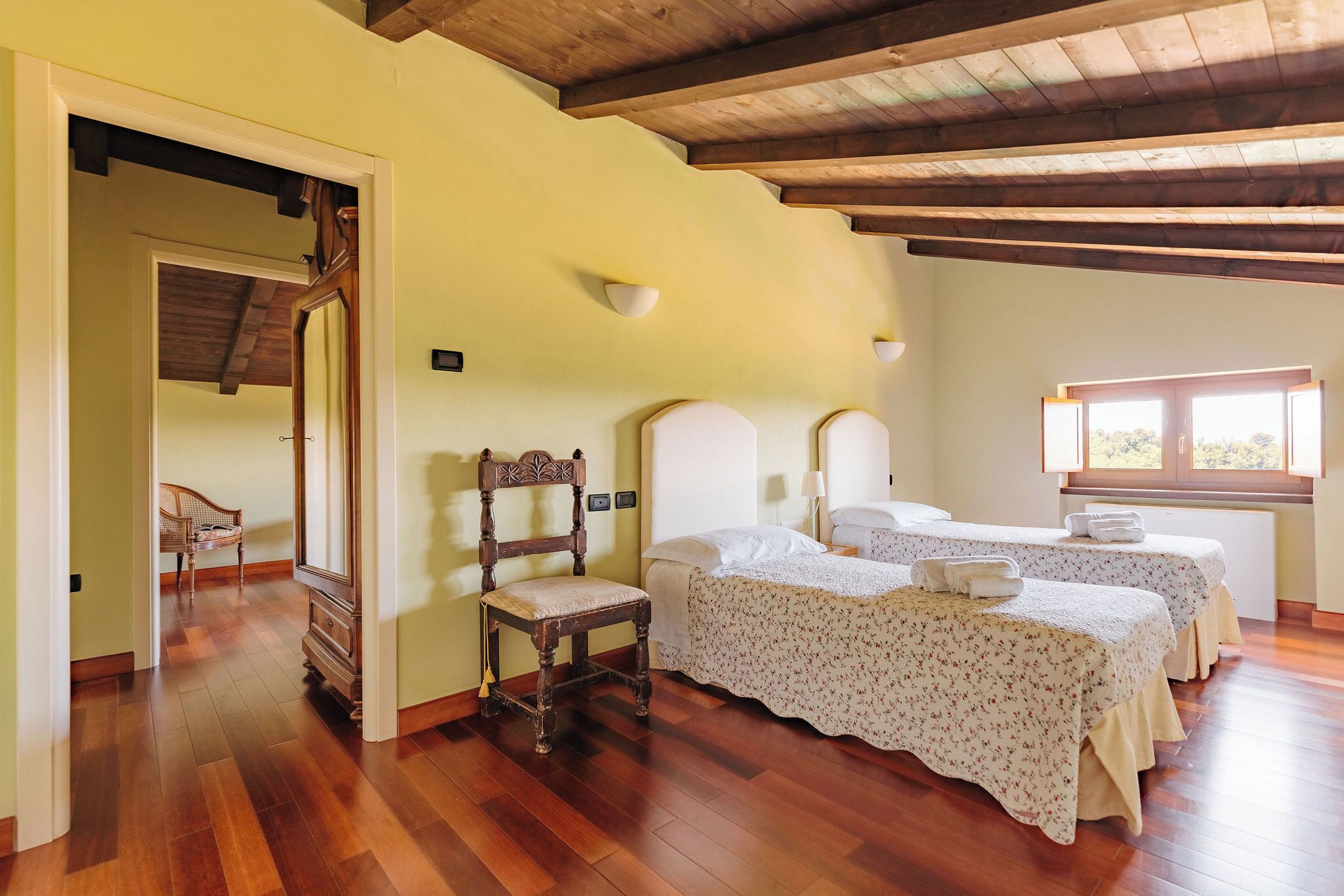 A room with two beds, wooden flooring, a chair, and a window.