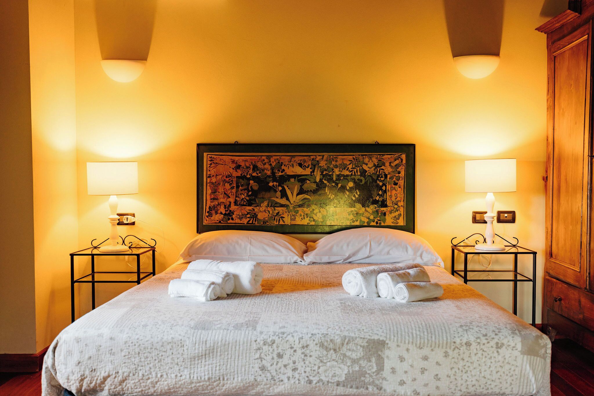 A hotel room with a large bed, two bedside lamps, a floral artwork above the bed, and a wooden wardrobe.