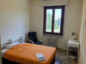 Quarto com cama, cadeira, escrivaninha, radiador e janela com vista para edifícios e árvores.