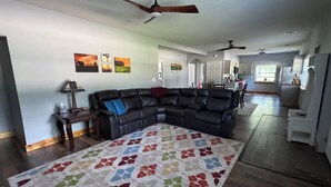 A spacious living room with a sectional sofa, a patterned rug, ceiling fans, and a kitchen in the background.