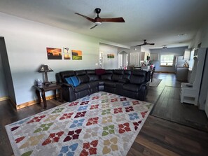A spacious living room with a sectional sofa, a patterned rug, ceiling fans, and a kitchen in the background.